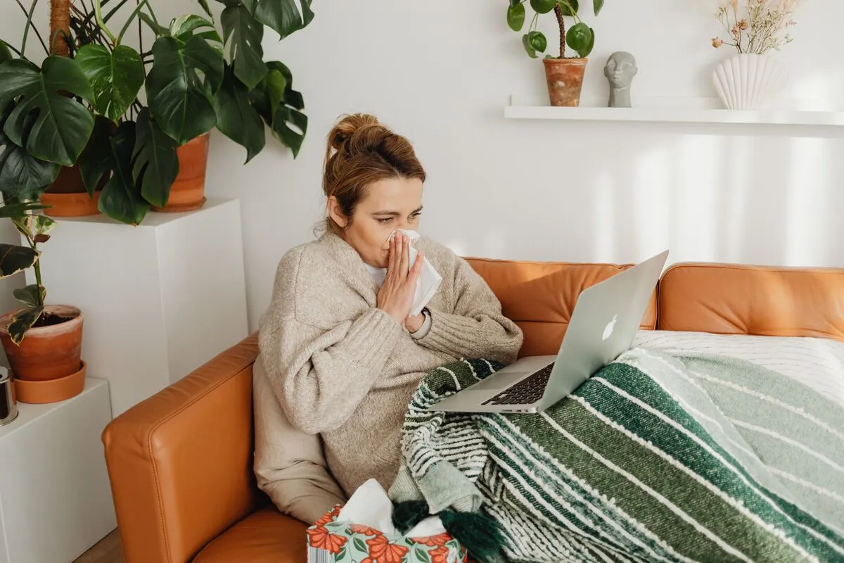 Voici des propositions de texte alternatif (attribut alt), adaptées selon l'objectif SEO et d'accessibilité de ton site : Option descriptive (recommandée pour l'accessibilité) : « Femme enrhumée sous un plaid sur un canapé, se mouchant avec un mouchoir tout en travaillant sur un ordinateur portable